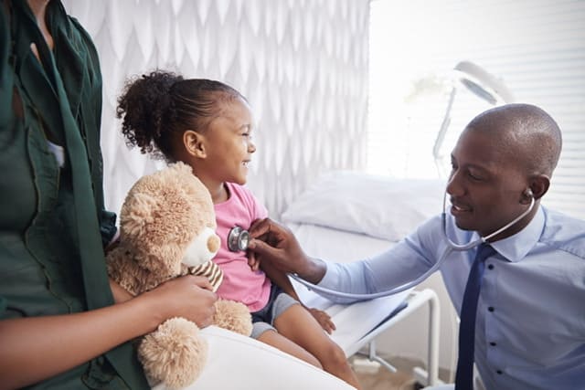 Black Doctor with stethoscope listening to the heartbeat of a black girl who is smiling. Her mom is sitting next to hear holding a teddy bear.