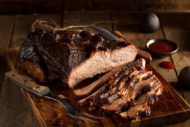 A beef brisket half sliced on a wooden cutting board with a carving knife and fork