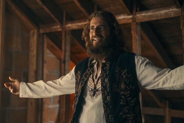 Hippie man standing with arms outstretched. Jonathan Roumie as Lonnie Frisbee in Jesus Revolution.