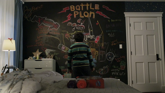 Boy with his back toward the camera looking at a chalkboard wall in a bedroom. The wall says Battle Plan and shows various ways of securing his household