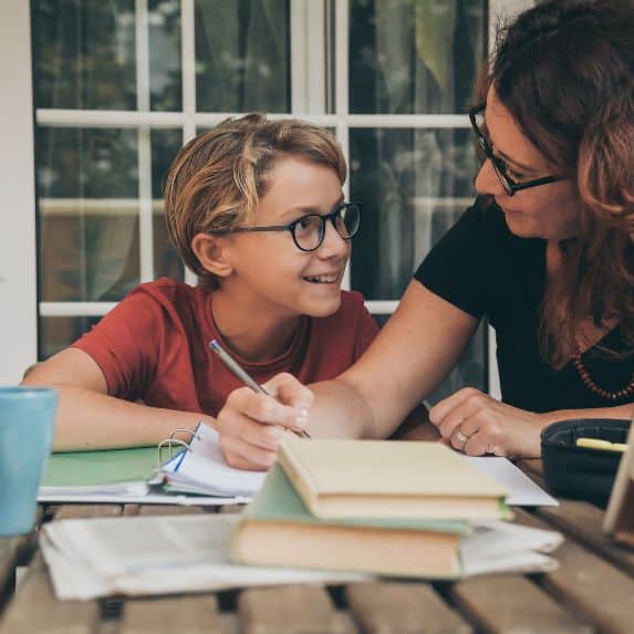 Mom with glasses and holding pen helping white boy with his schoolwork. Homeschooling