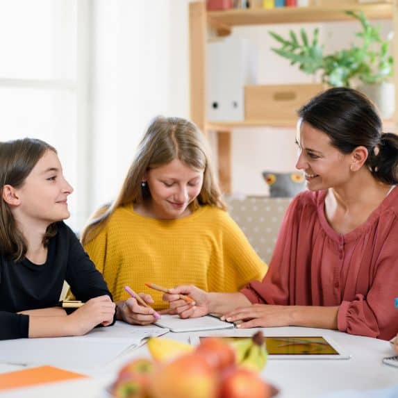 A woman sitting on the kitchen table with two girls working on school.