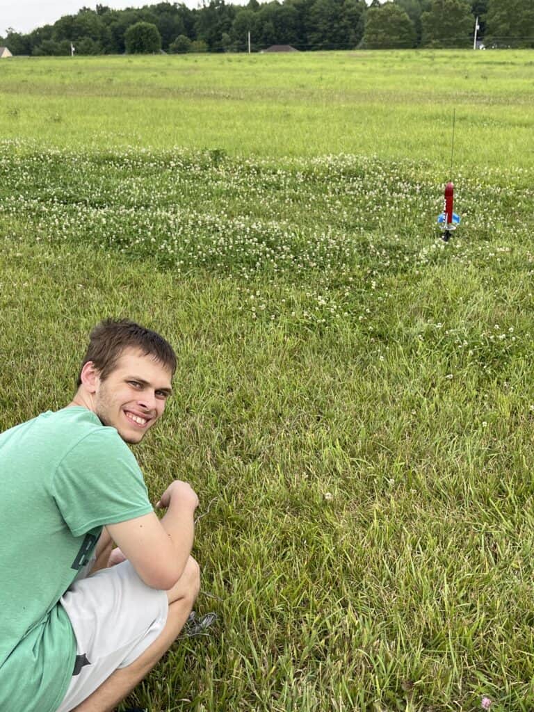 Boy crouched down in a field of green grass and clover with a red model rocket in the background waiting to launch.