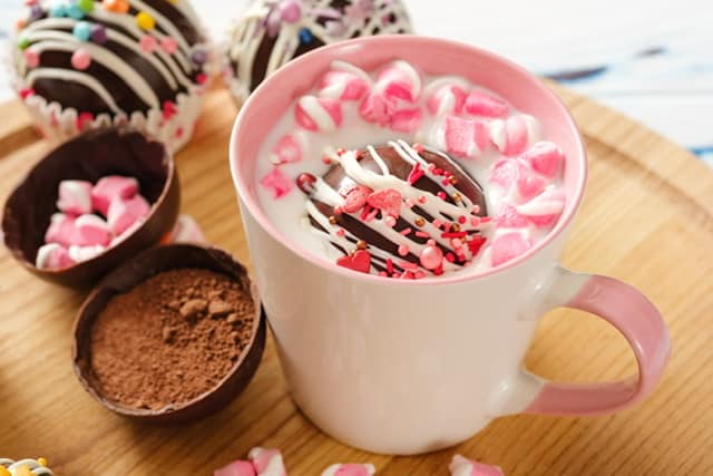 cup of milk with hot chocolate bomb sitting on a wood board surrounded by pink and white candies and chocolate bombs