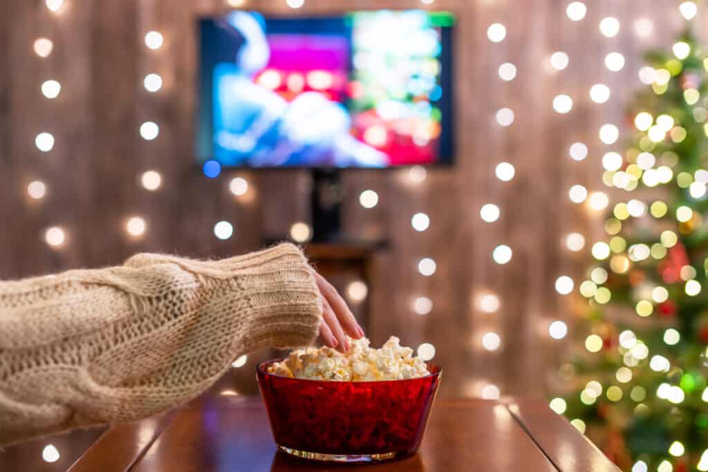 End table with red glass bowl filled with popcorn. A hand is reaching into the bowl. In the background there is the twinkle of Christmas lights and a movie playing on the television.