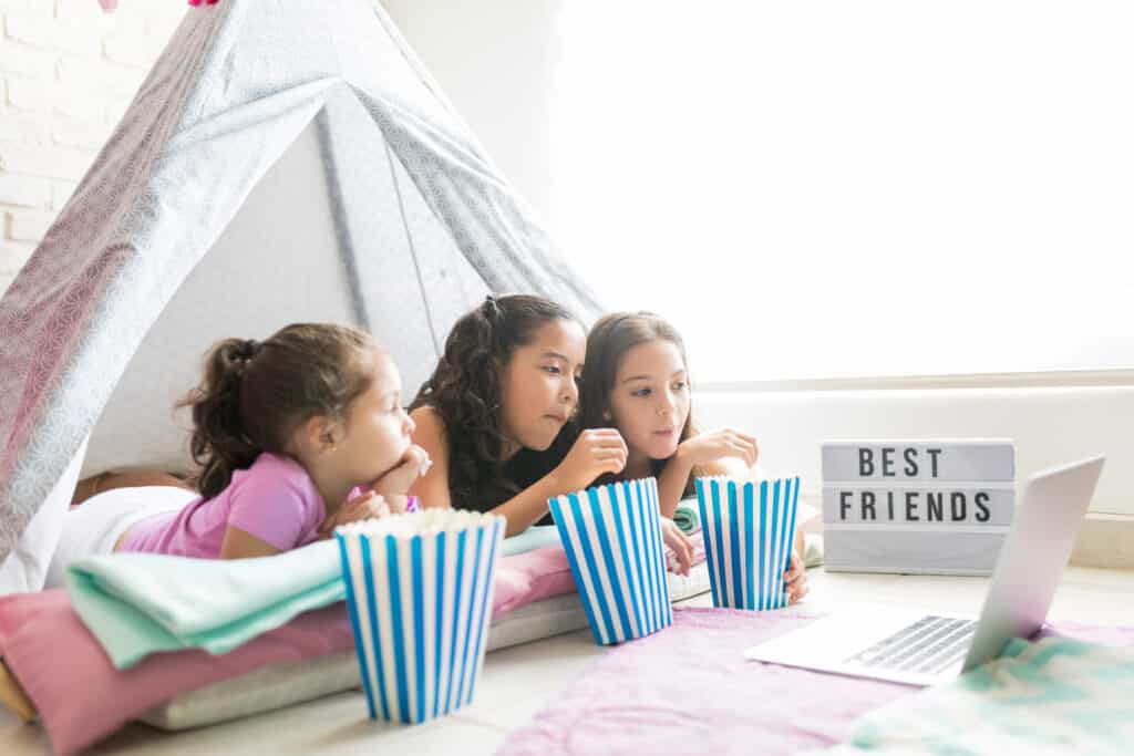Best Friends - three girls watching a movie on a laptop. They each have blue and white stripped popcorn buckets and a inside a tent.