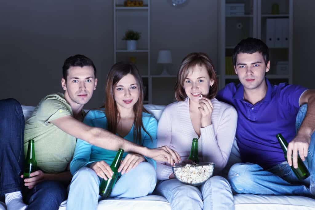 Two couples drinking beer out of bottled and watching a movie while eating popcorn.