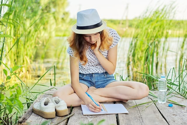 Girl sitting and writing on a dock near the water with tall grasses around her.
