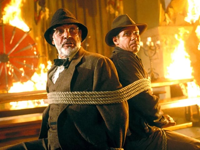 Sean Connery as Henry Jones Sr, and Harrison Ford as Indiana Jones tied together in chairs with a fire burning around theml.