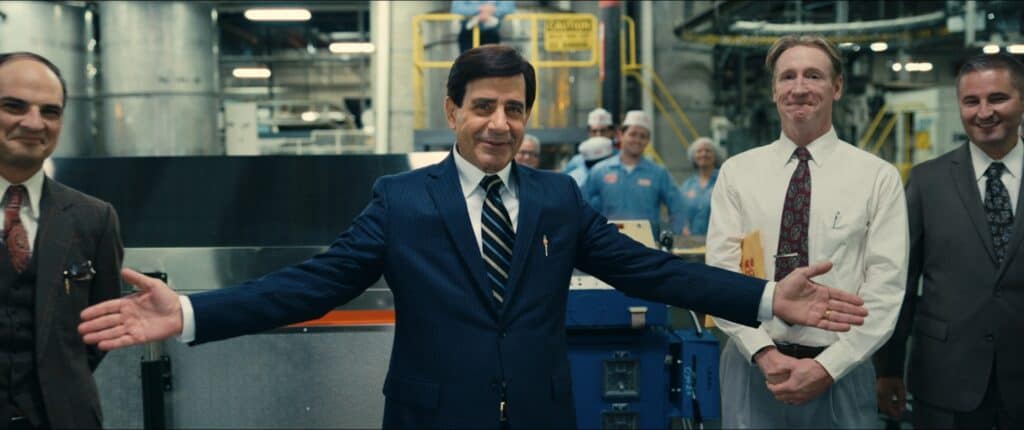 A Middle Aged white man with black hair and wearing a navy blue suit and striped tie has his arms outstretched and welcoming. He stands in a factory with other white man in suits and ties.