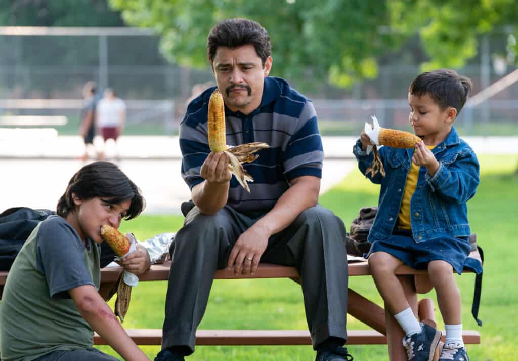 A Mexican American Man and two boys sit on picnic table eating Mexican street corn.