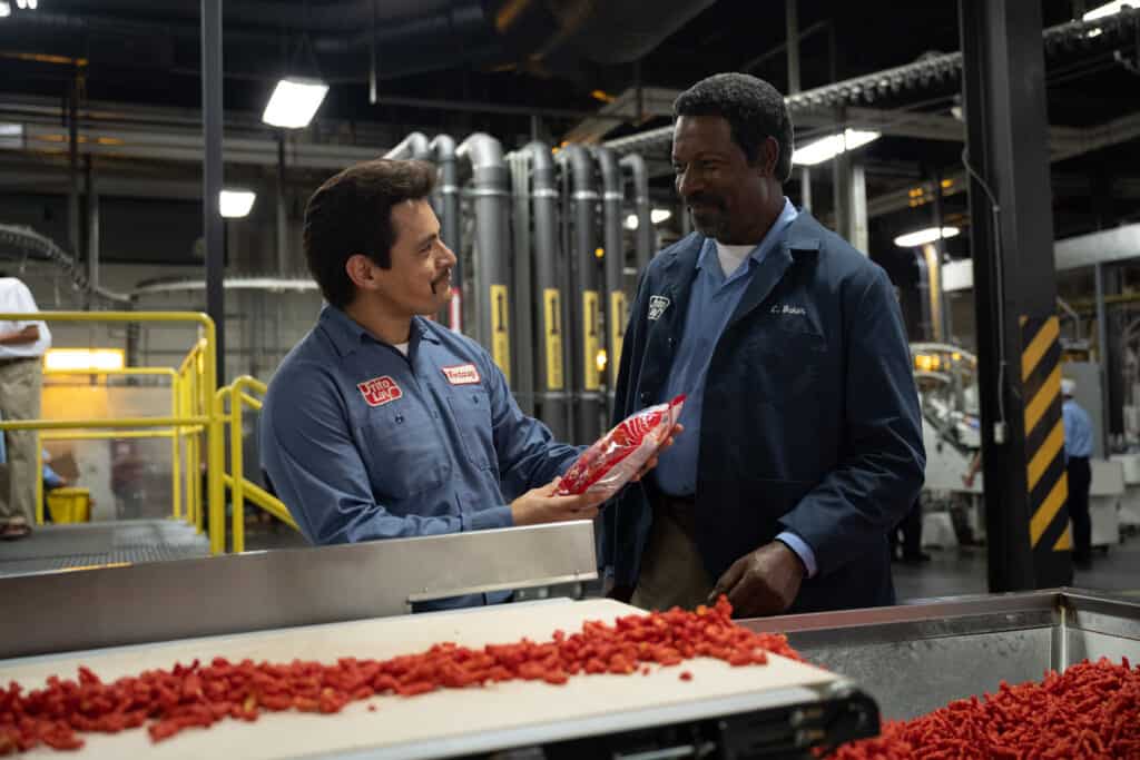 A Mexican-American man and a Black man standing in a Frito-Lay factory with Flamin' Hot Cheetos.