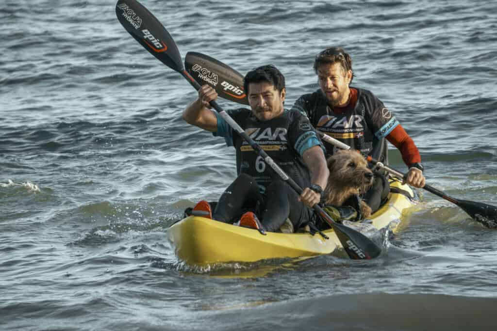2 men in a yellow canoe paddling with a dog in the middle of the canoe. Simu Liu as Leo and Mark Wahlberg as Michael in Arthur the King.
