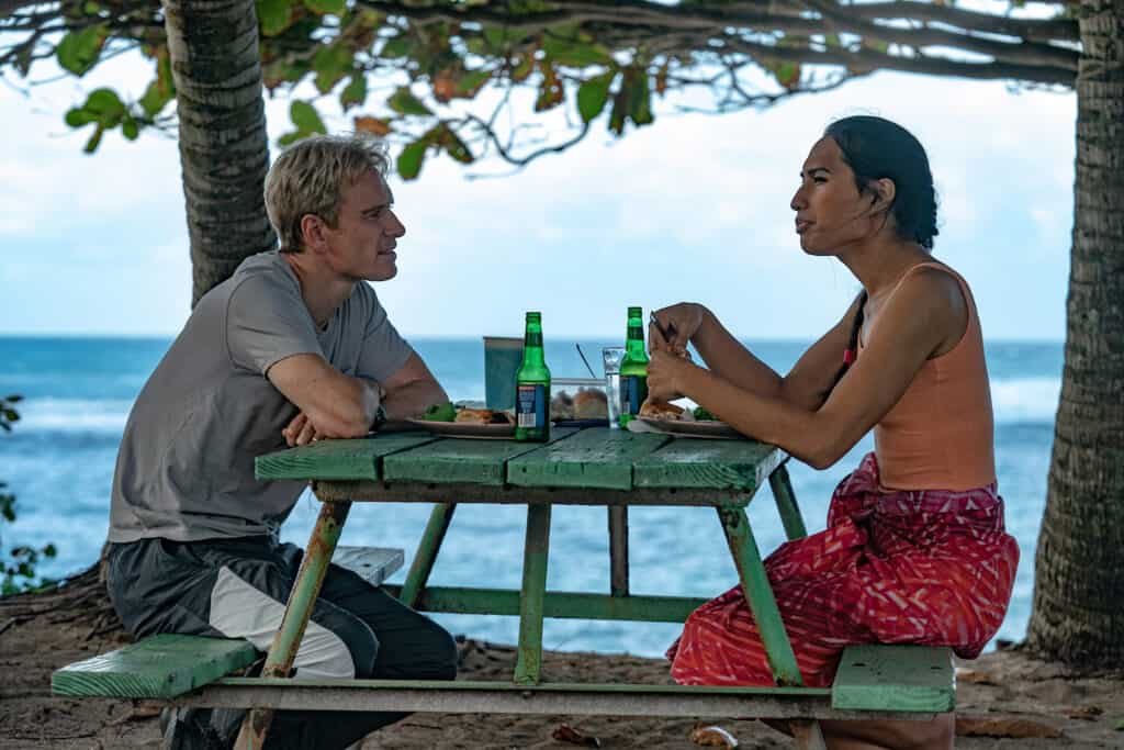 A blond man and a dark skinned woman sit on a picnic bench drinking beers and eating lunch. They are overlooking the ocean.