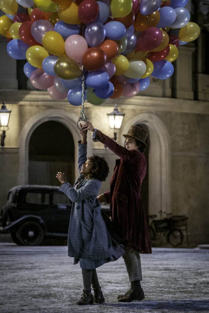 A young man in a tophat and a black girl hold onto a bunch of balloons at night in the town center.