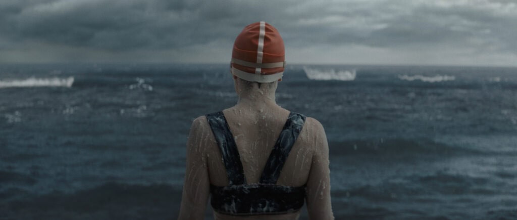 Swimmer with her back toward the screen. She is wearing an orange swimming cap and looking out over the sea.